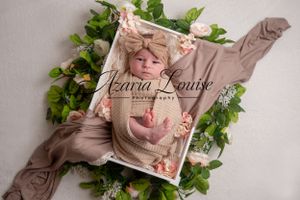 Newborn photography featuring a sleeping baby wrapped in tan fabric with greenery accents and wooden crate.