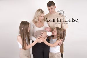 Family of four in light colored clothing gathers around newborn in studio photo shoot.