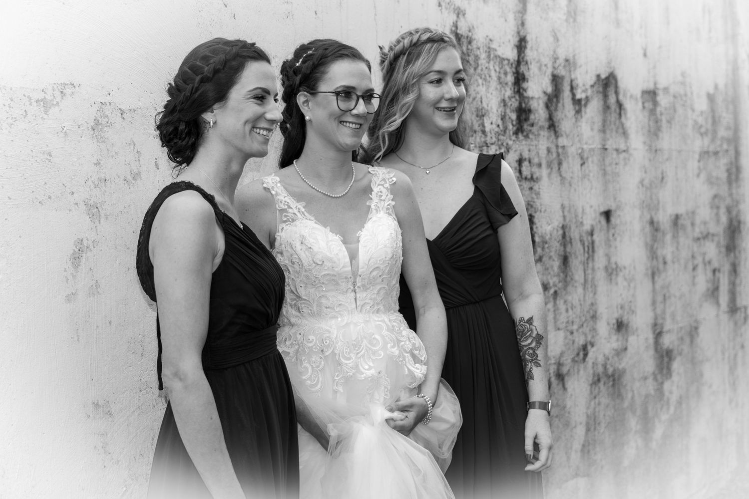 Bride in lace gown smiles with two bridesmaids in black dresses in black and white photo.
