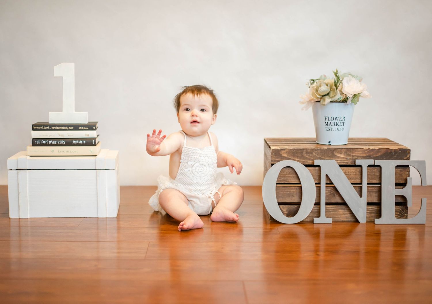 Baby sitting on wooden floor with decorative 'ONE' letters and books in background.