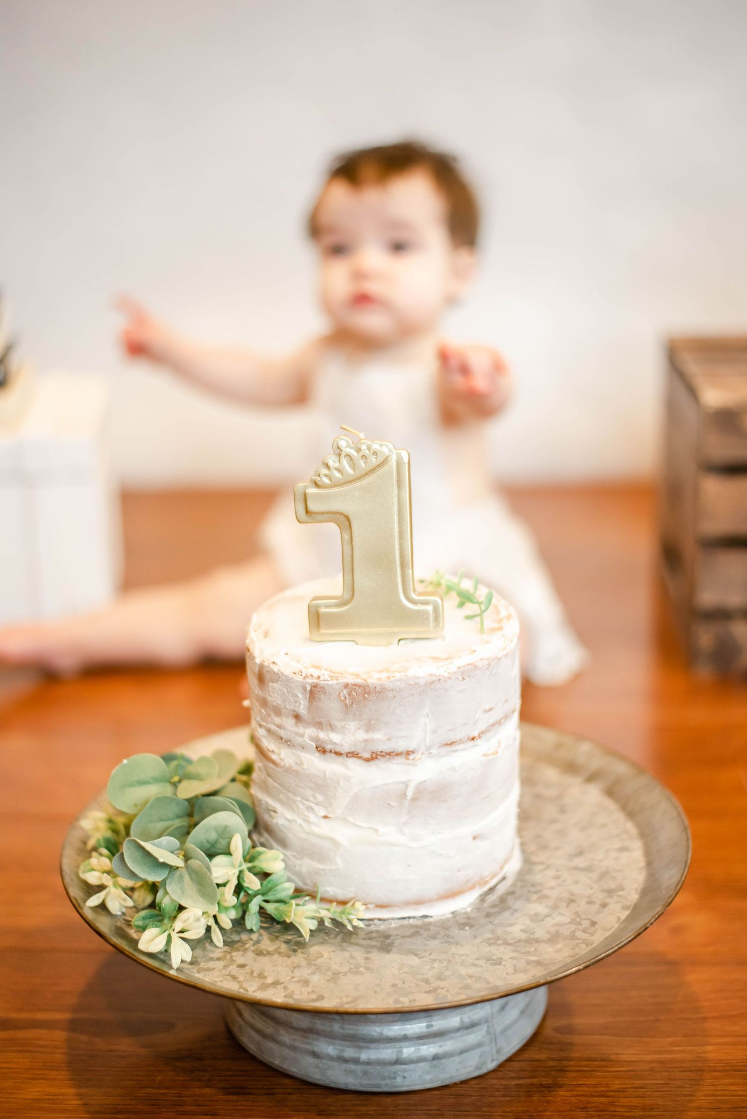 White birthday cake with gold number one topper and greenery on metal cake stand.