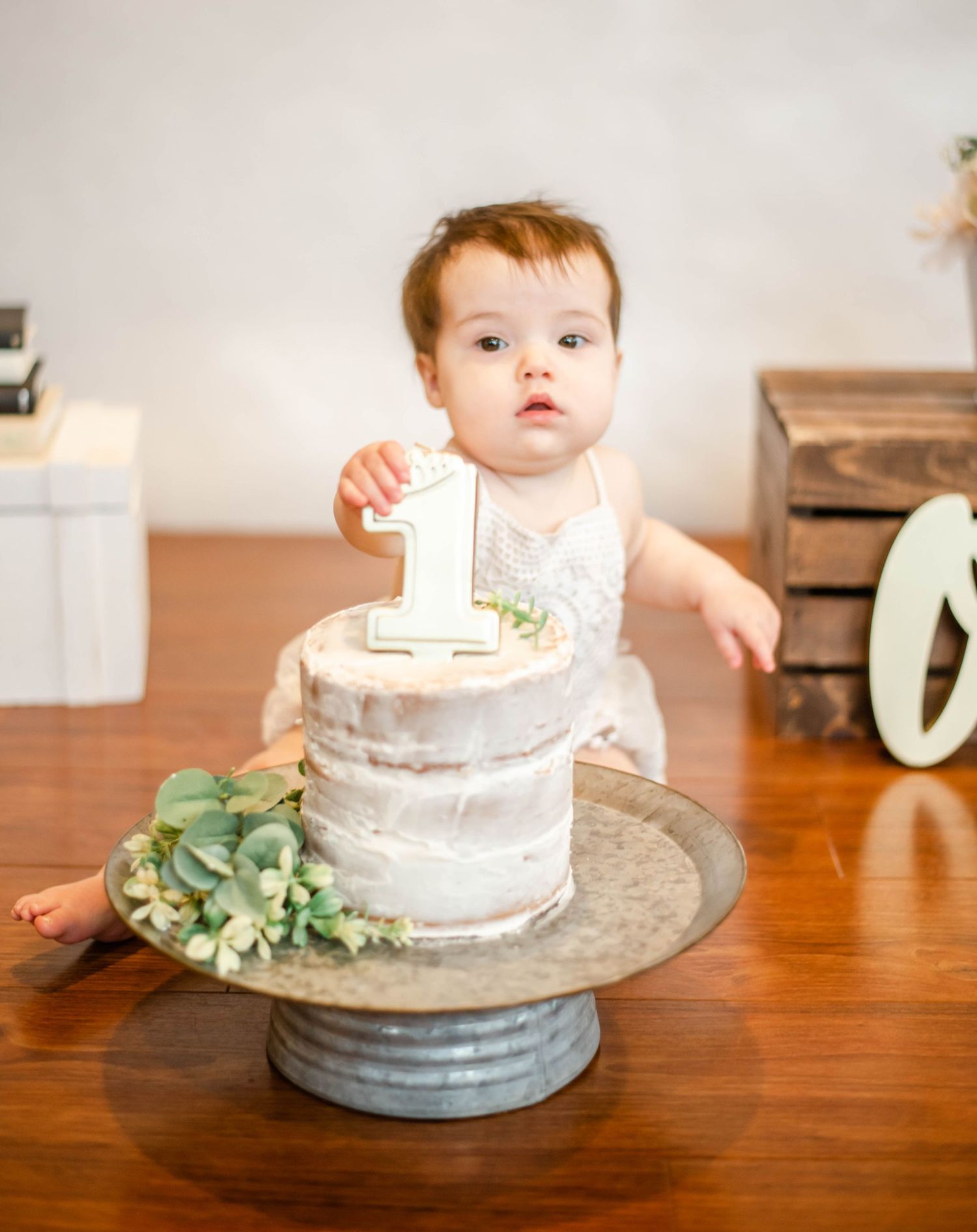 A simple white cake sits on a gray platter atop a wooden table with first birthday decorations.