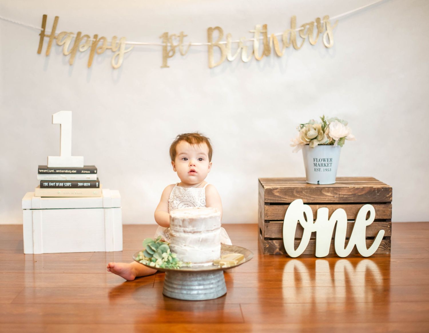 Birthday setup with a white cake and gold banner reading 'one' against a clean white wall.
