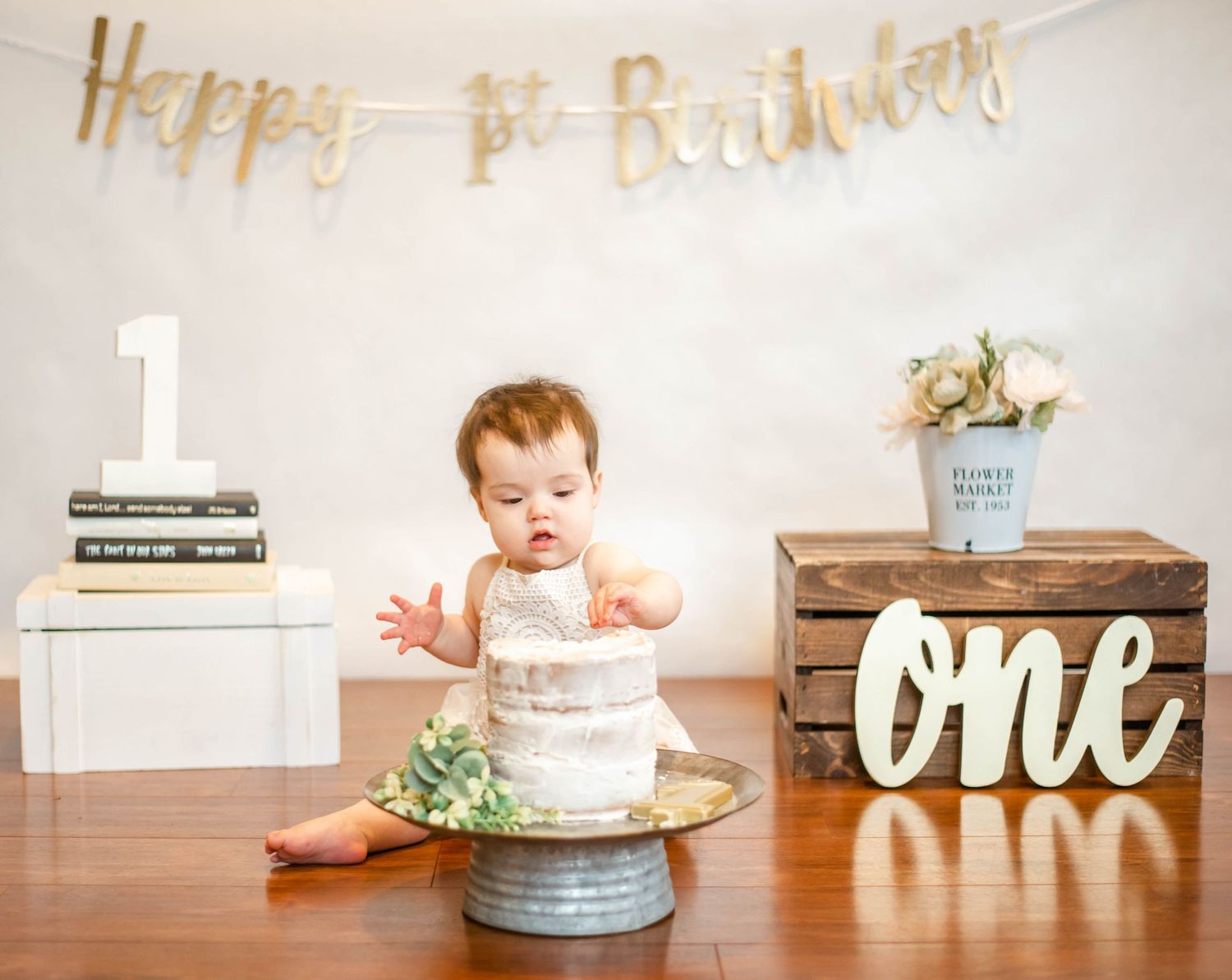 White smash cake setup with rustic decorations and gold 'one' sign for first birthday celebration.