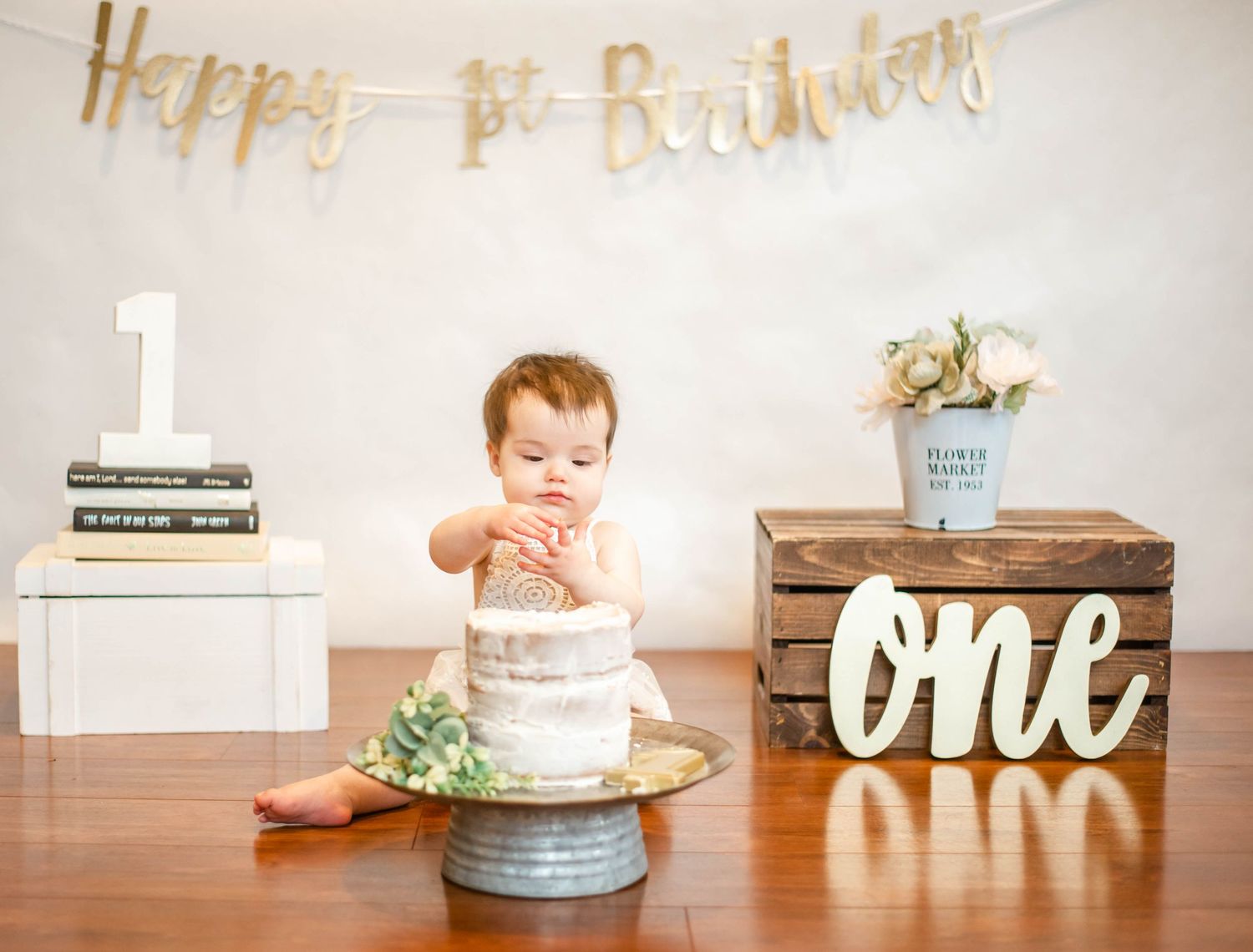 First birthday party scene with white cake and golden banner decorations on wooden surface.
