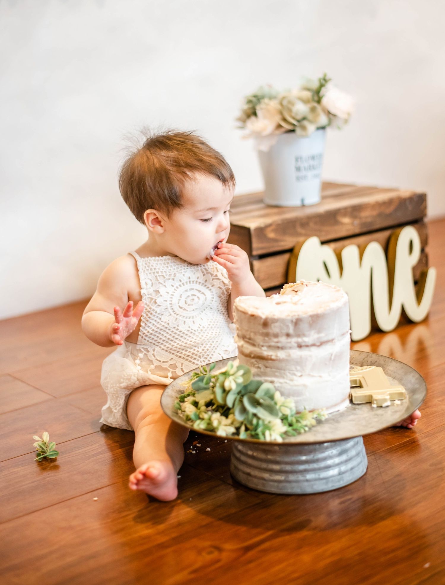 Baby reaches for white tiered cake topped with greenery on wooden table in rustic setting.