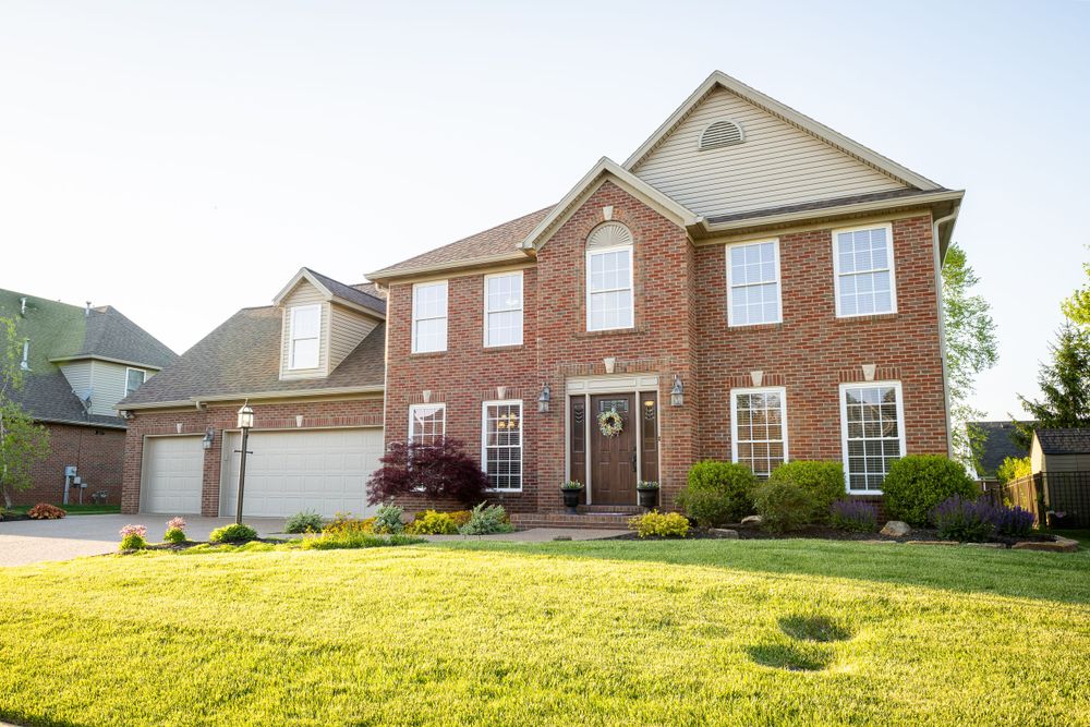 Large two-story brick colonial home with attached garage and manicured lawn in a suburban setting.