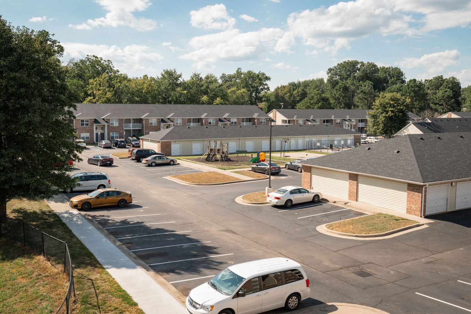 Aerial view of a residential apartment complex with white buildings and paved parking areas under a partly cloudy sky.