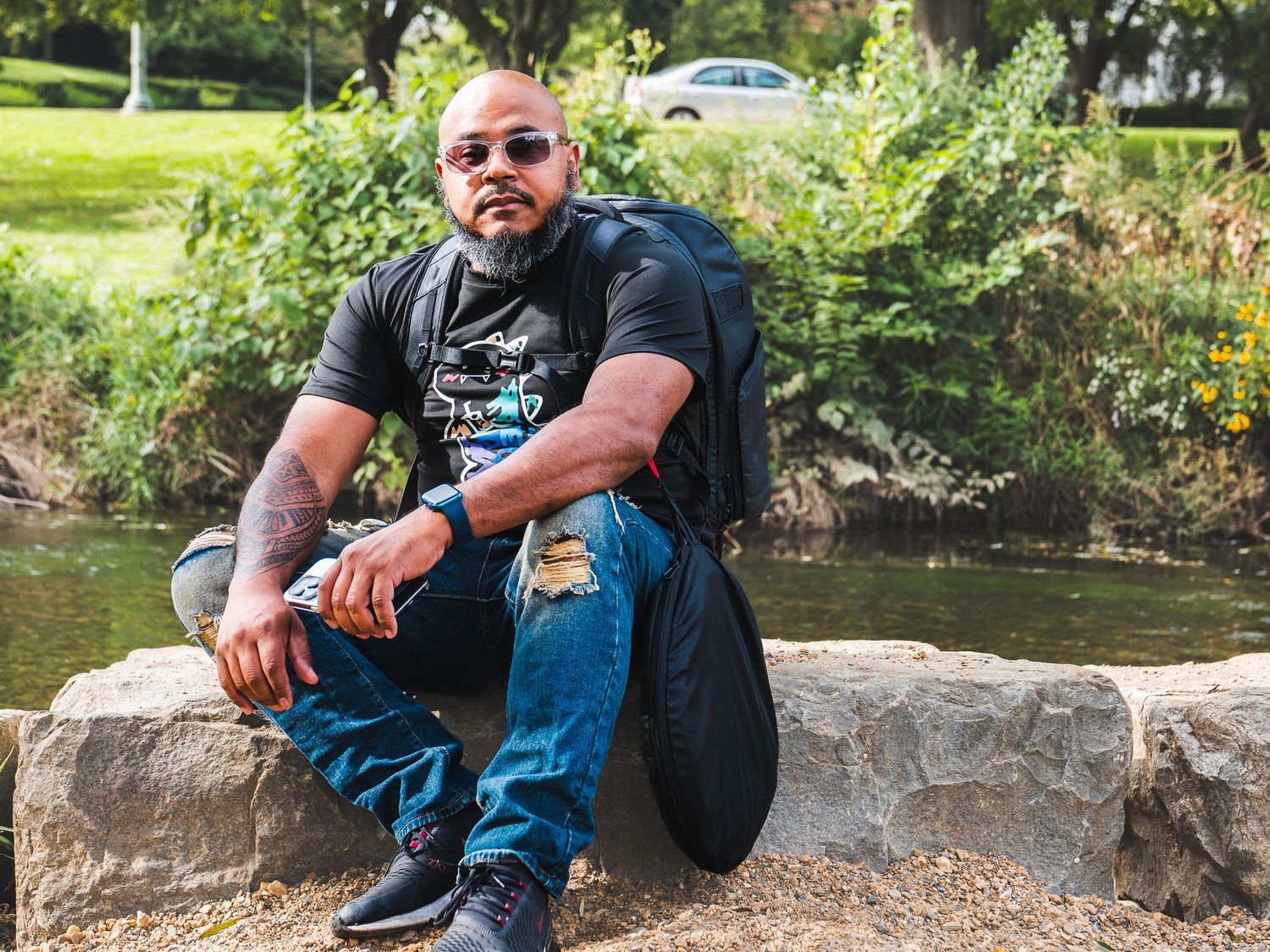 A man in sunglasses sits casually on a rock by a stream wearing casual clothing and a backpack.