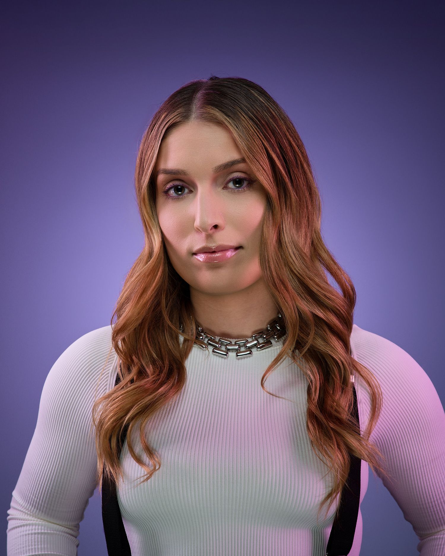 Professional portrait with long wavy auburn hair wearing a white ribbed sweater and layered necklace against purple background.