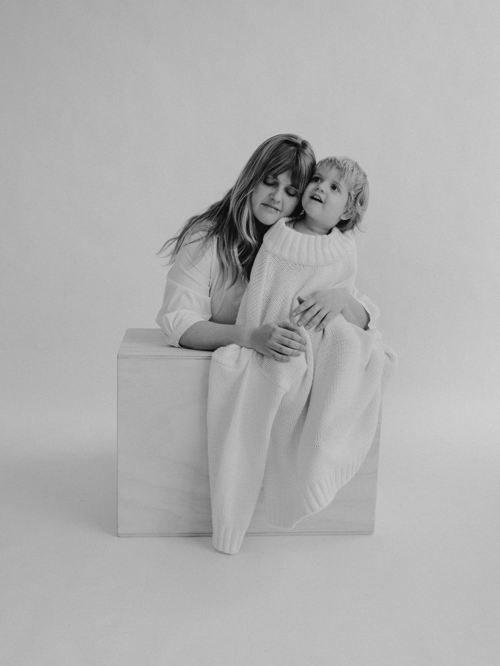 Retrato en estudio fotográfico en blanco y negro de una madre con su hijo que viste un jersey muy grande de punto blanco.