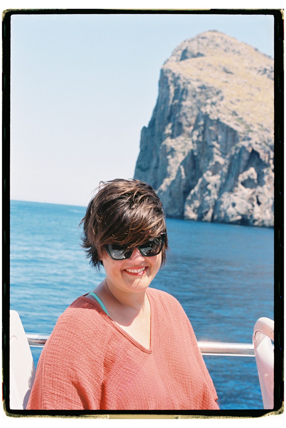 Person in coral sweater smiling on boat with coastal cliff in background.