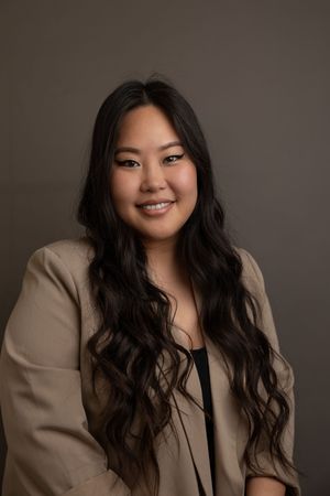 Professional business headshot of a smiling woman wearing a black top and beige jacket with studio lighting against a dark grey backdrop.