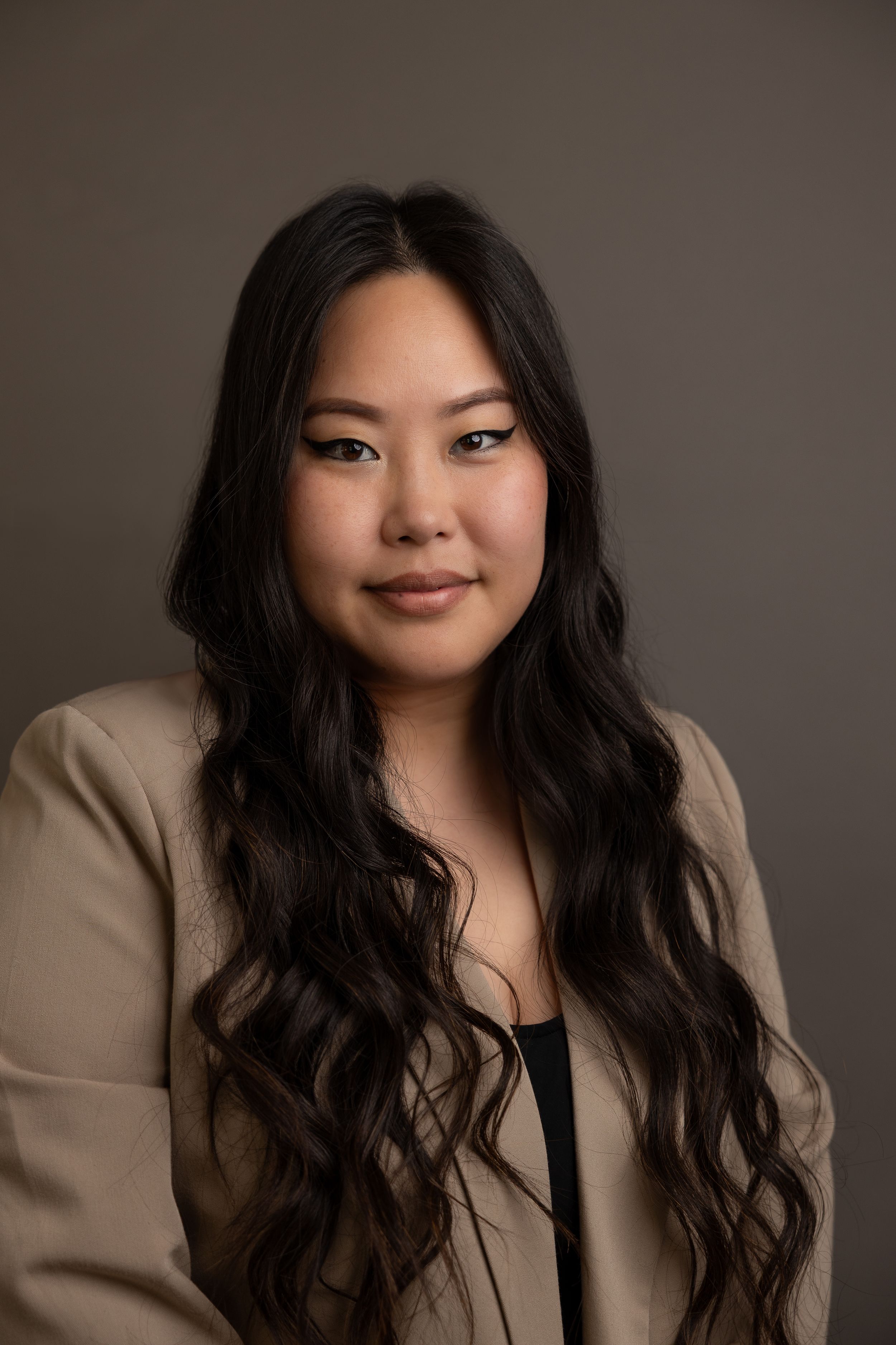 Professional business headshot of a woman wearing a black top and beige jacket with studio lighting against a dark grey backdrop.