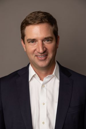 Professional business headshot of man wearing a white shirt and dark blazer with studio lighting against a dark grey backdrop.