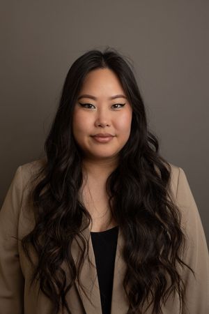 Professional business headshot of a woman wearing a black top and beige jacket with studio lighting against a dark grey backdrop.