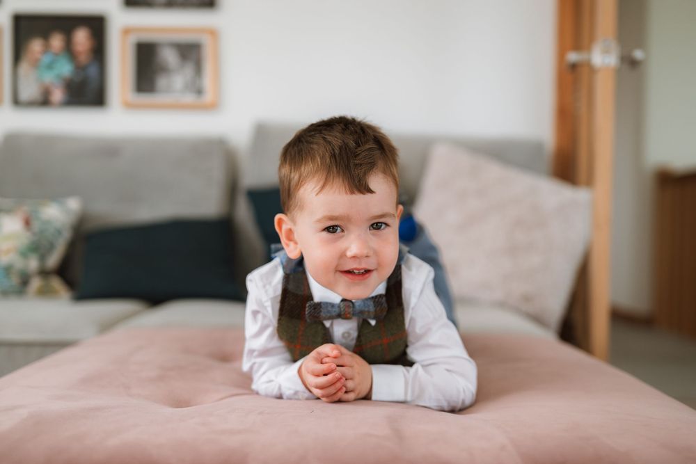 A young child in formal attire lies on a couch smiling while propped up on their elbows.