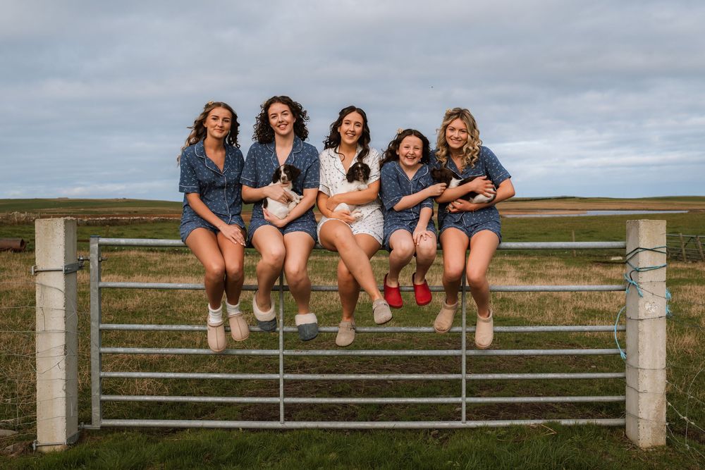 A group sits together on a metal farm gate wearing denim jackets and boots against a cloudy rural landscape.