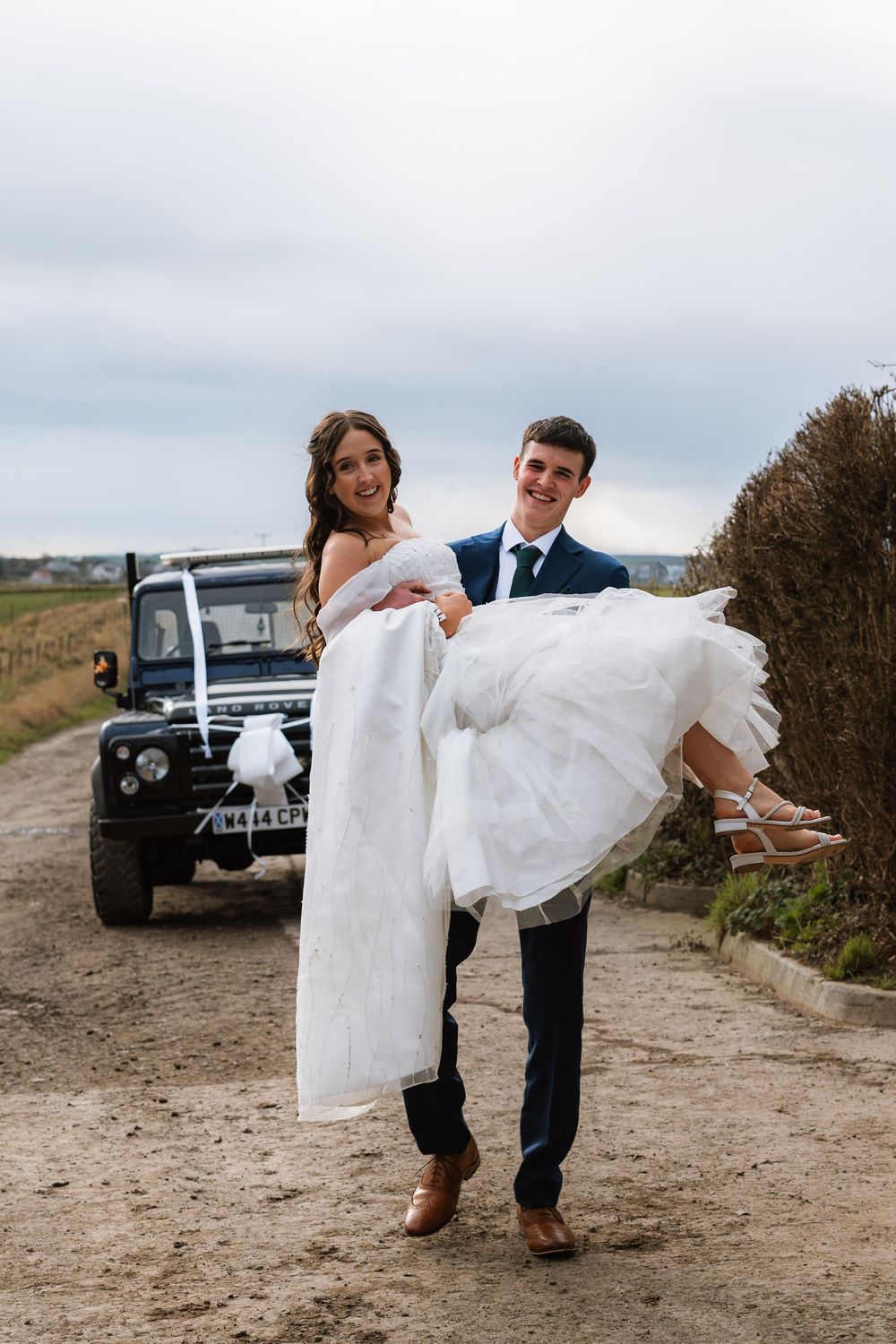 A newlywed couple poses playfully in front of a vintage Land Rover on a country path.