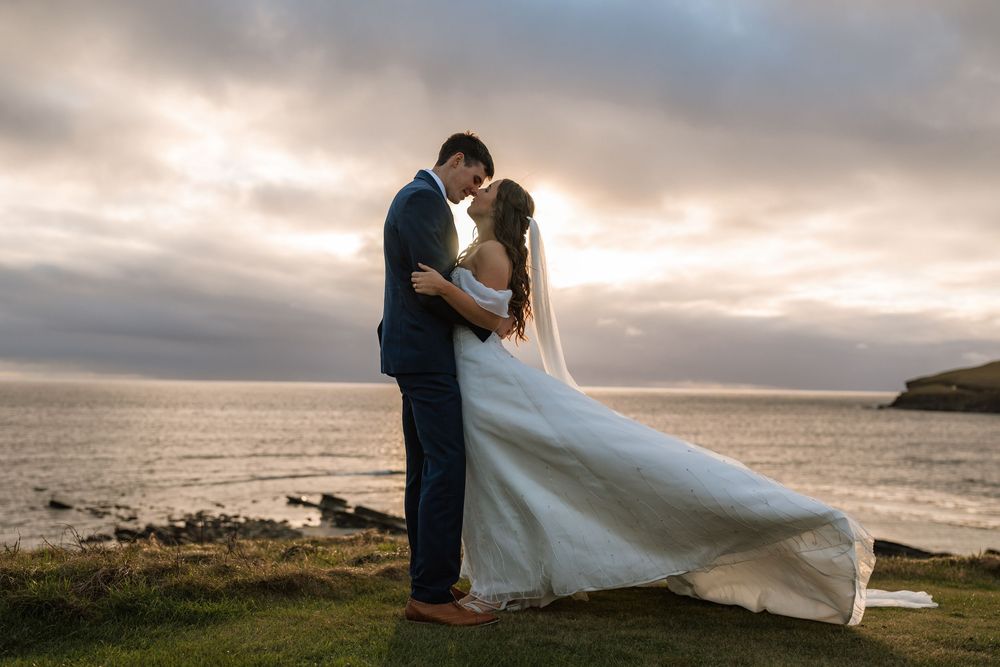 A romantic wedding couple embraces on a coastal cliff at sunset while the bride's dress flows in the wind.