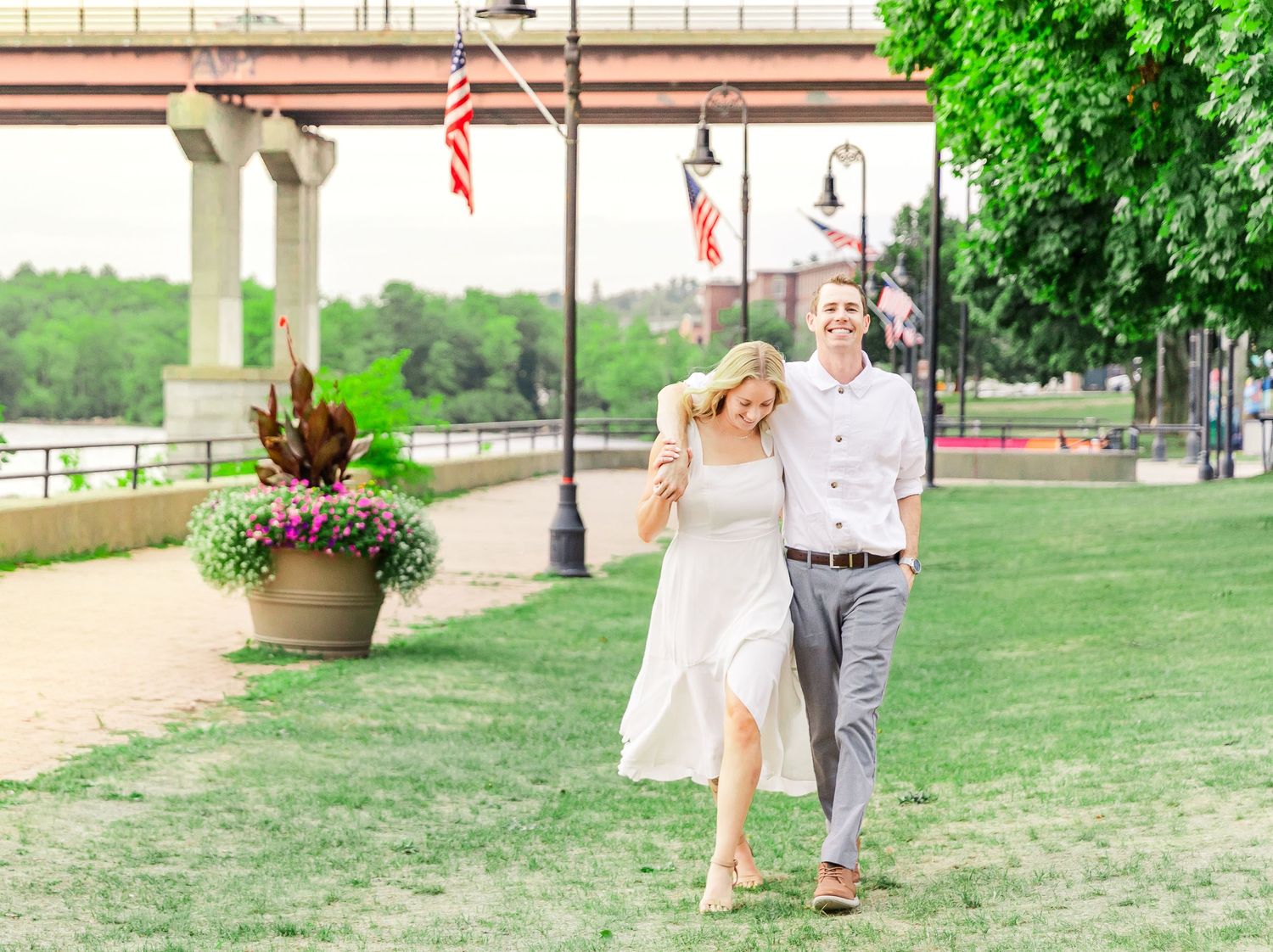Couple walks along riverside path with American flags and landscaped planters.