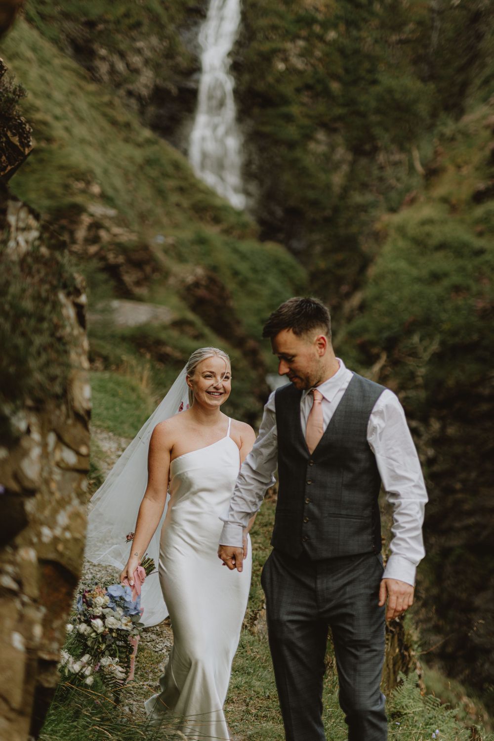 Couple poses together on path with dramatic waterfall backdrop in nature setting.