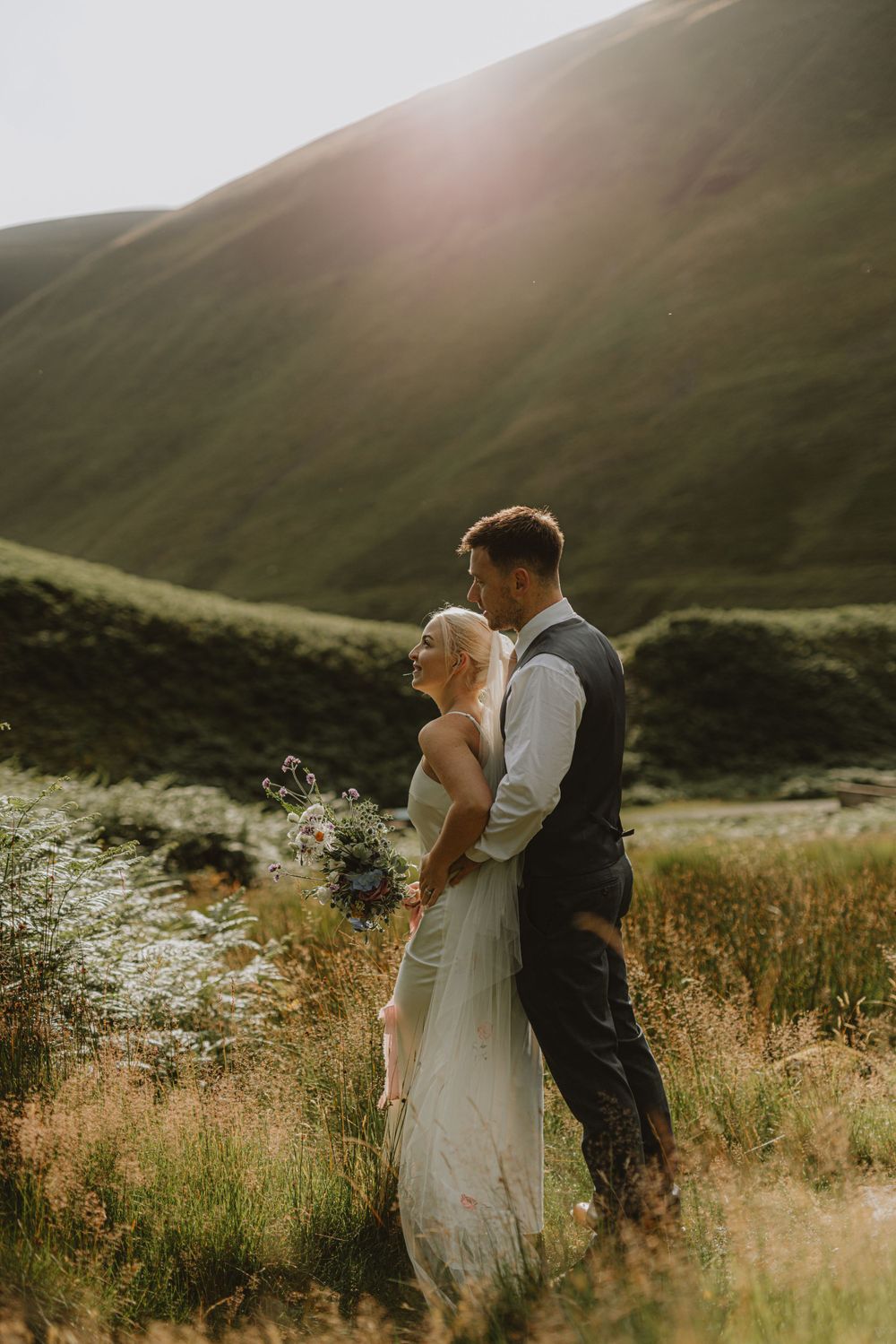 Newlyweds share an intimate moment against a backdrop of sunlit rolling hills and tall grass.