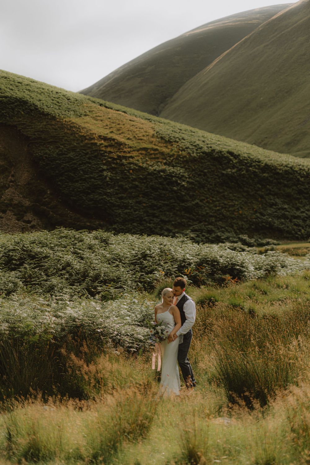 Wedding couple embraces in a scenic mountain valley with golden grass and green slopes.