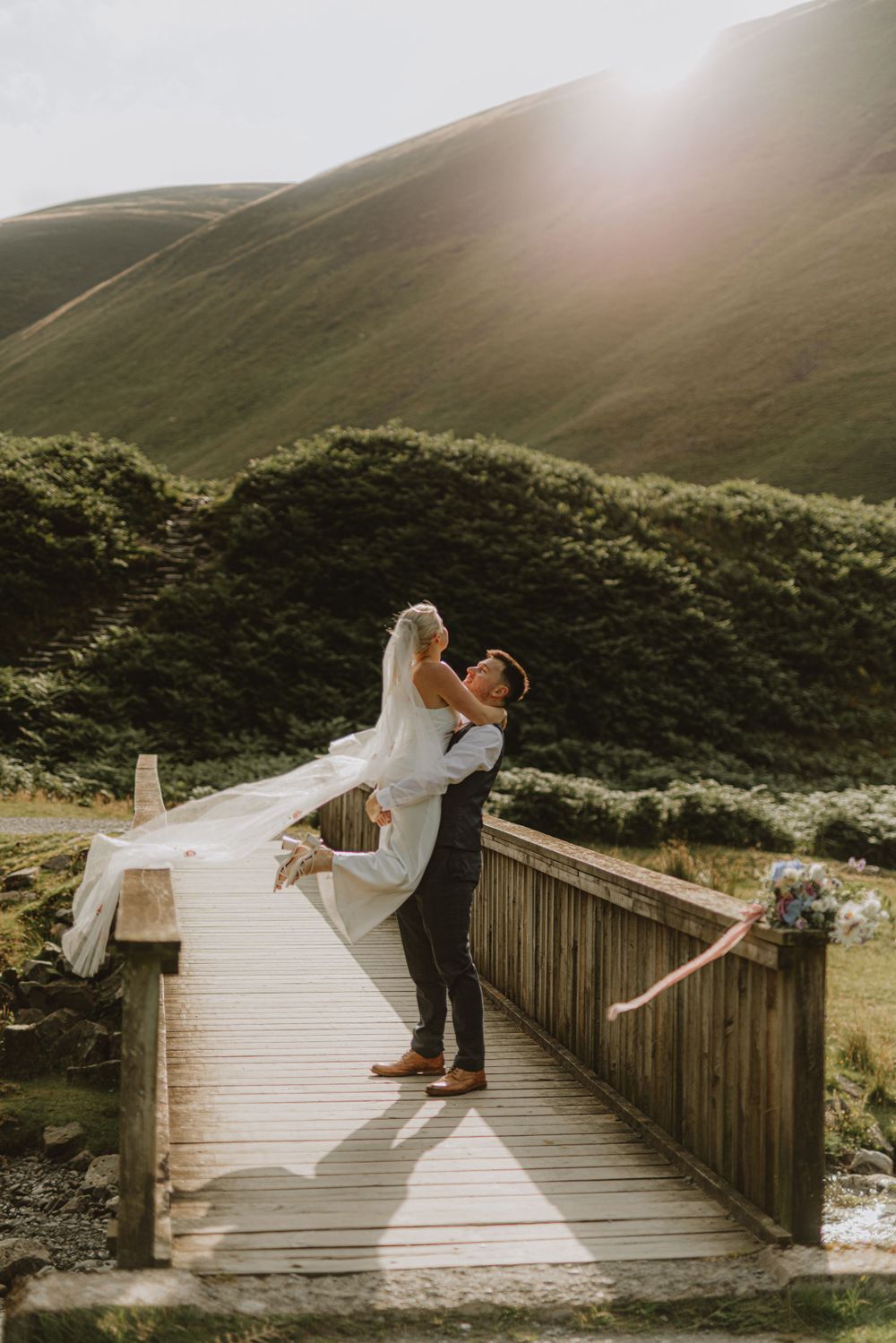 Wedding couple poses dramatically on a wooden bridge with mountains in the background.