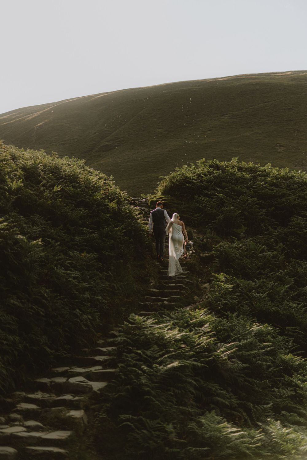 A couple ascends stone steps surrounded by lush greenery in a mountainous landscape.