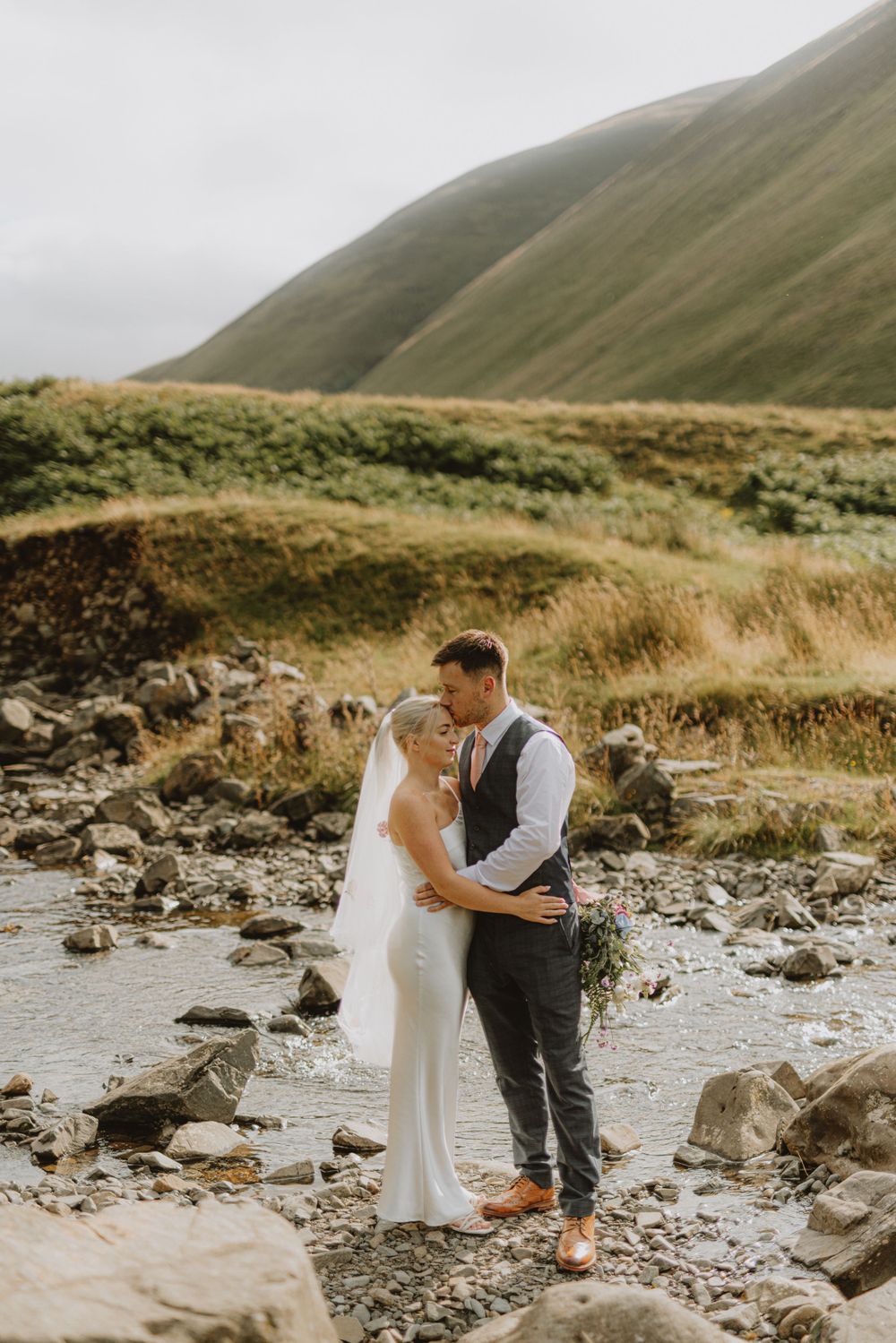 Wedding couple shares a tender moment on a rocky riverbank surrounded by mountains.