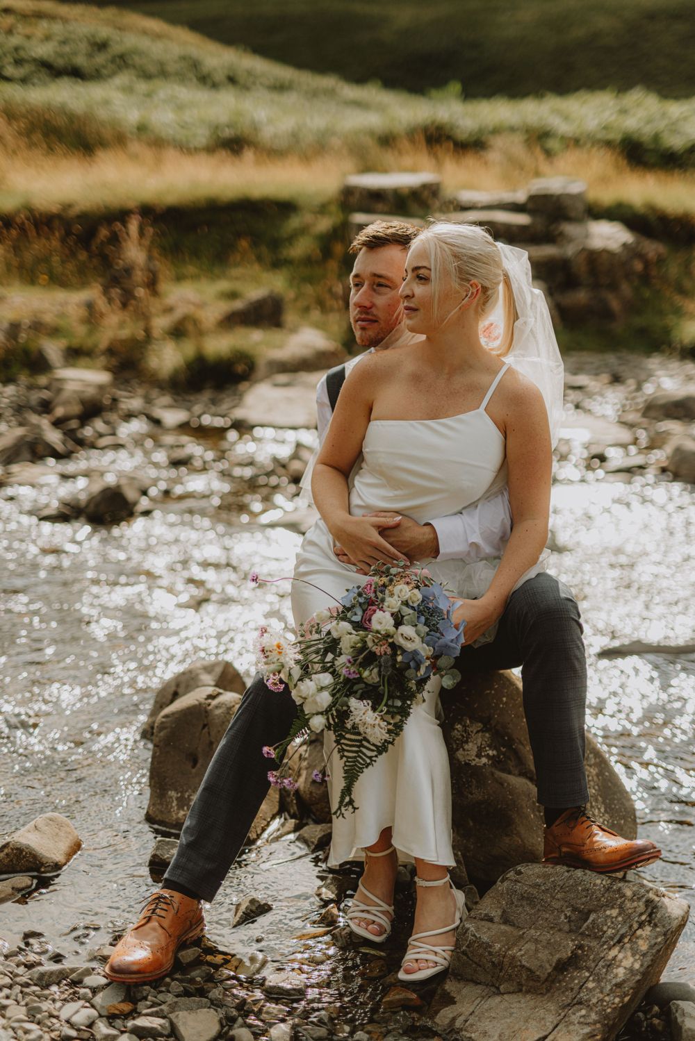 Couple sits intimately on rocks by the water holding a purple and white bouquet.