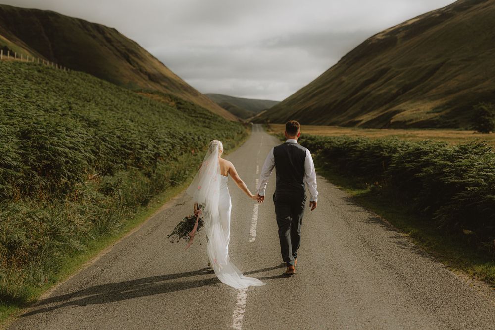 Newlyweds walk hand in hand down a scenic mountain road at their destination wedding.