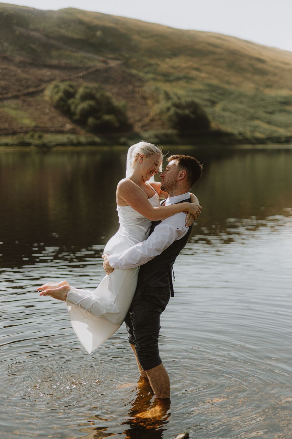 A romantic wedding couple embraces while standing in a serene lake with mountains in the background.
