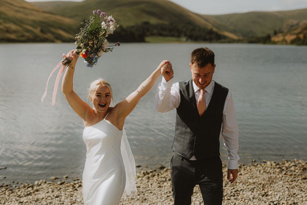 A joyful wedding celebration by a scenic lake with rolling hills in the background.