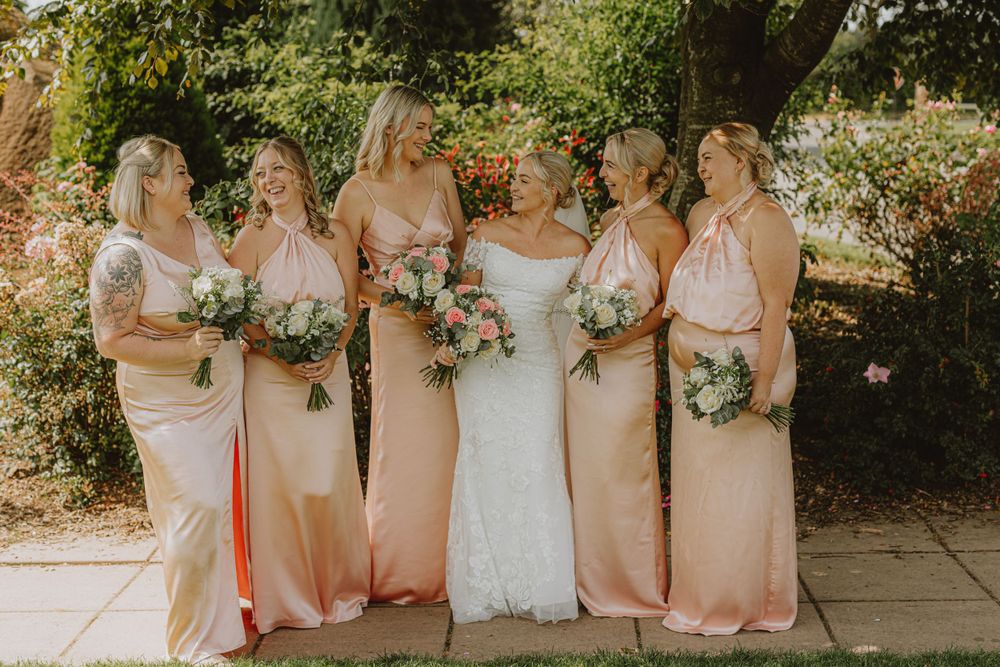 A bridal party in blush pink dresses poses together in a garden setting.