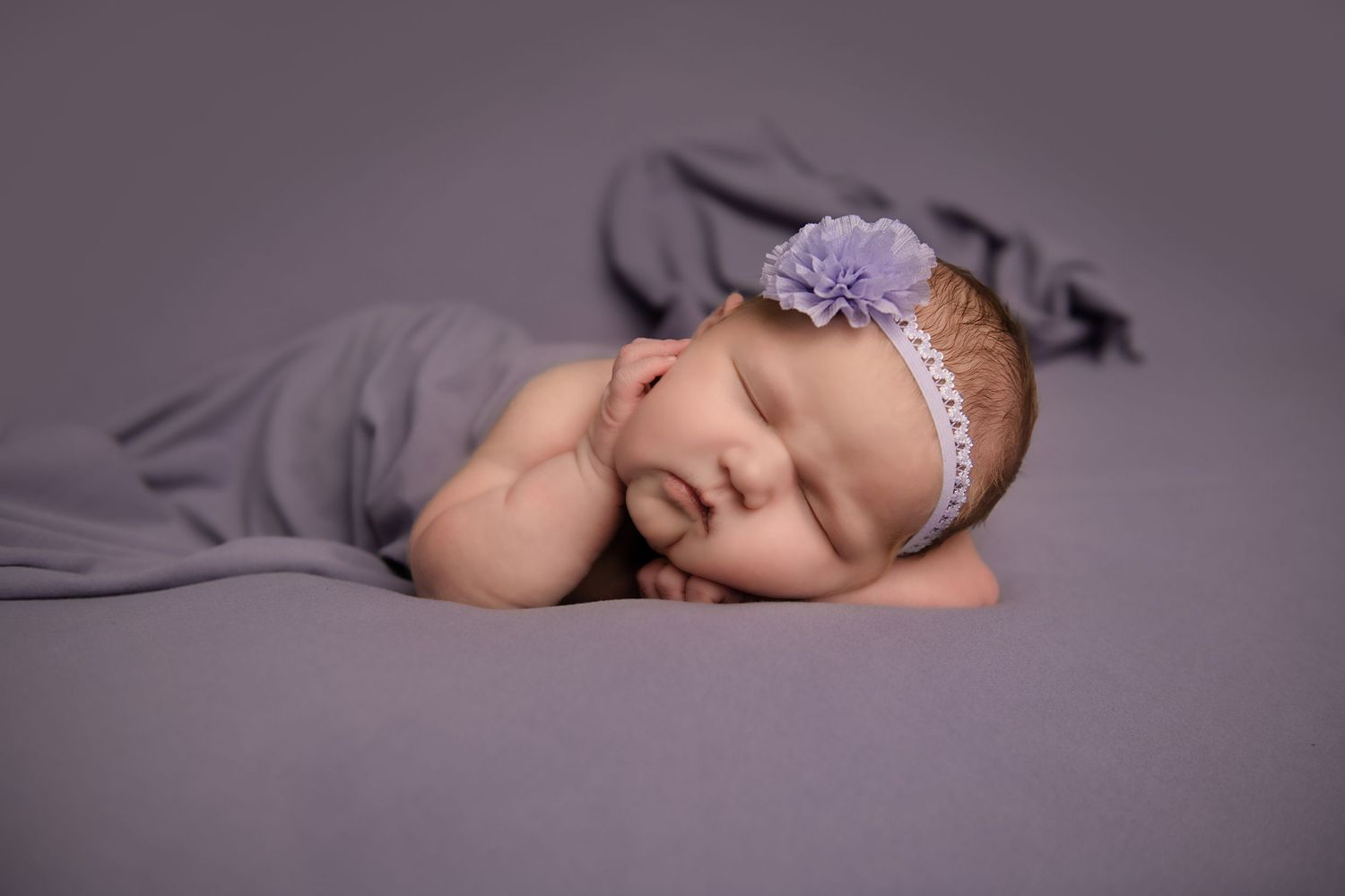 Sleeping newborn wearing purple headband on gray blanket during studio photoshoot.