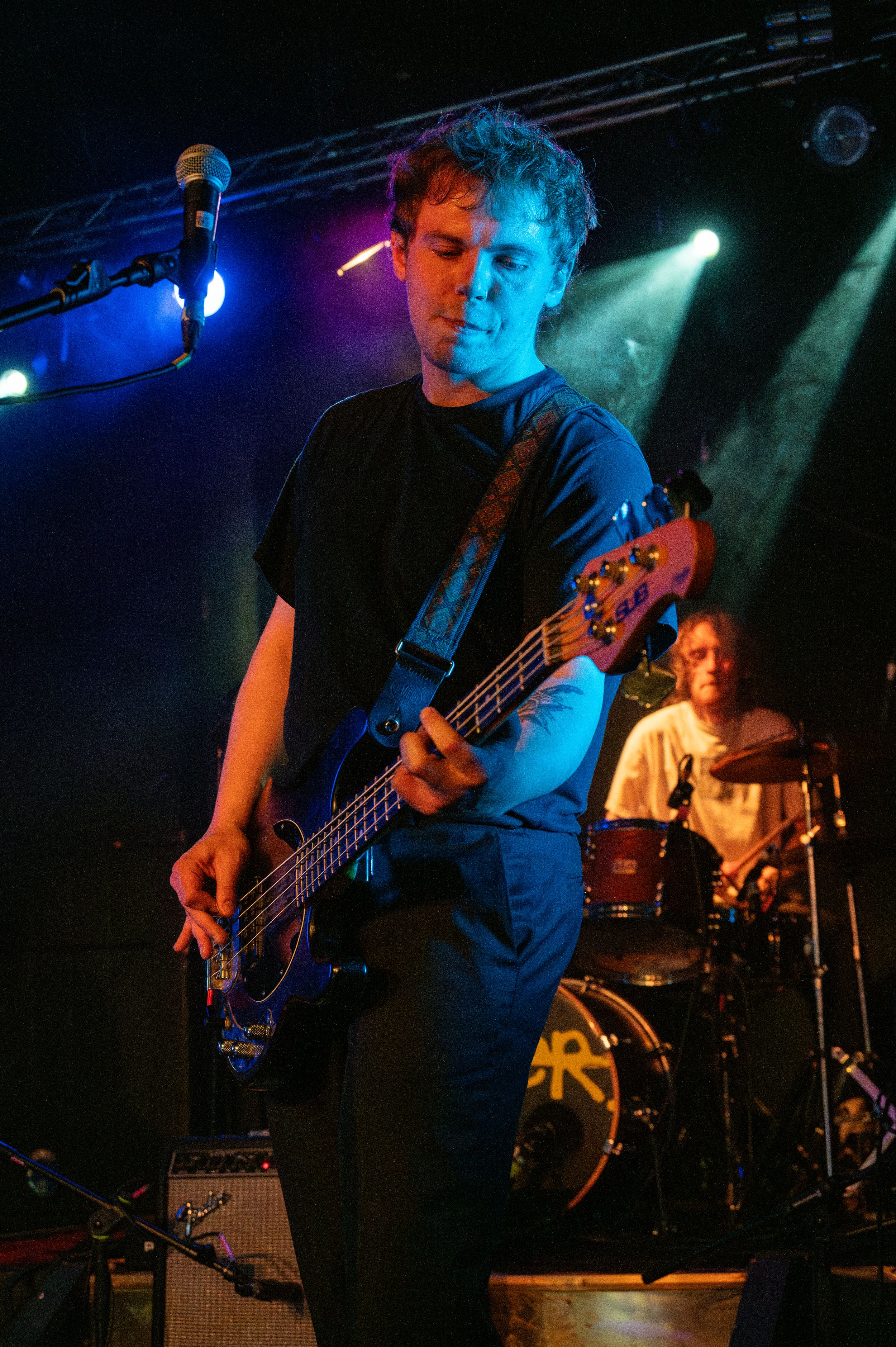 Bass guitarist performs on stage under dramatic blue and green lighting during a live concert performance.