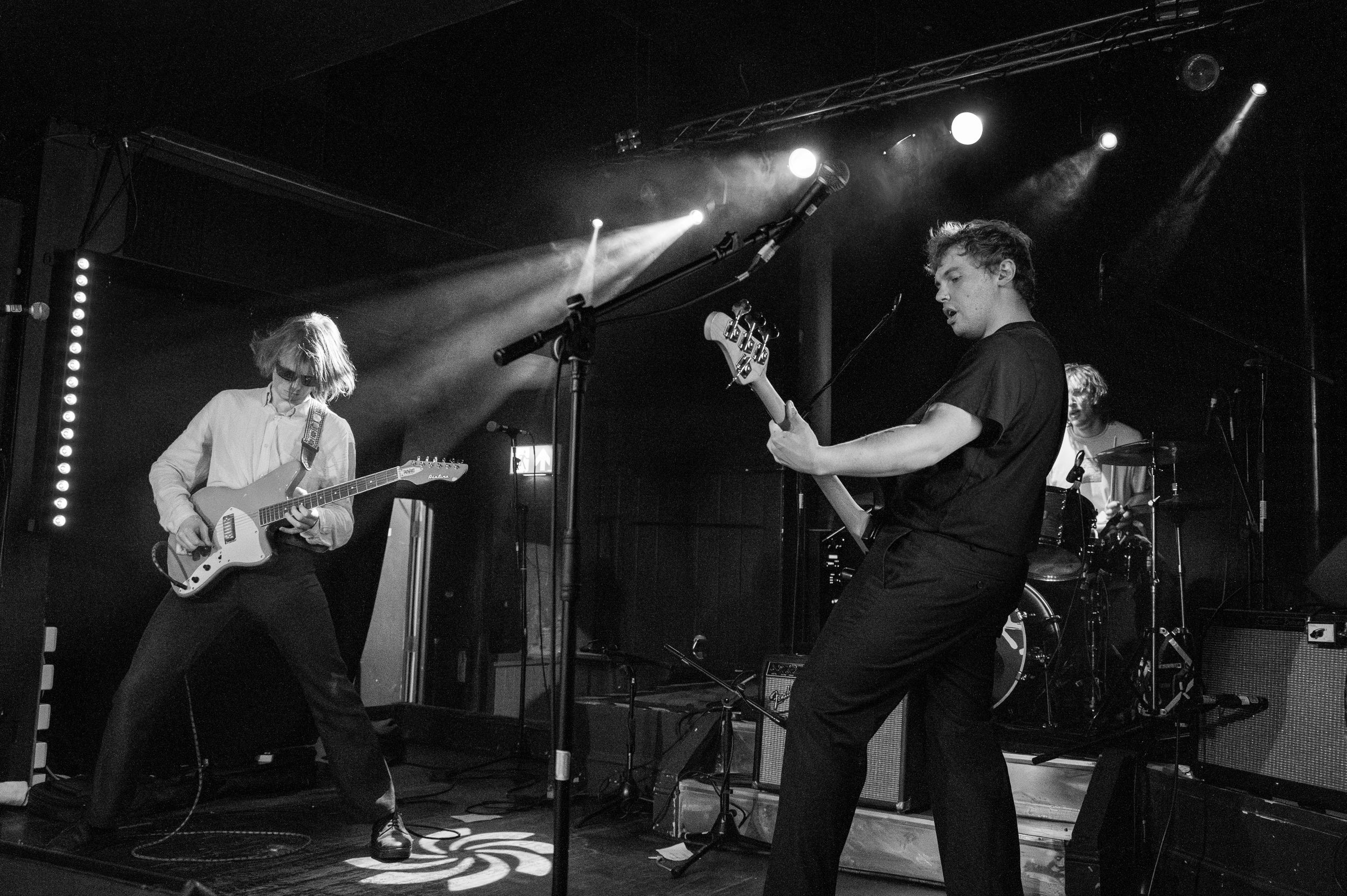 Black and white shot of guitar players performing energetically on stage with drum kit visible in background.