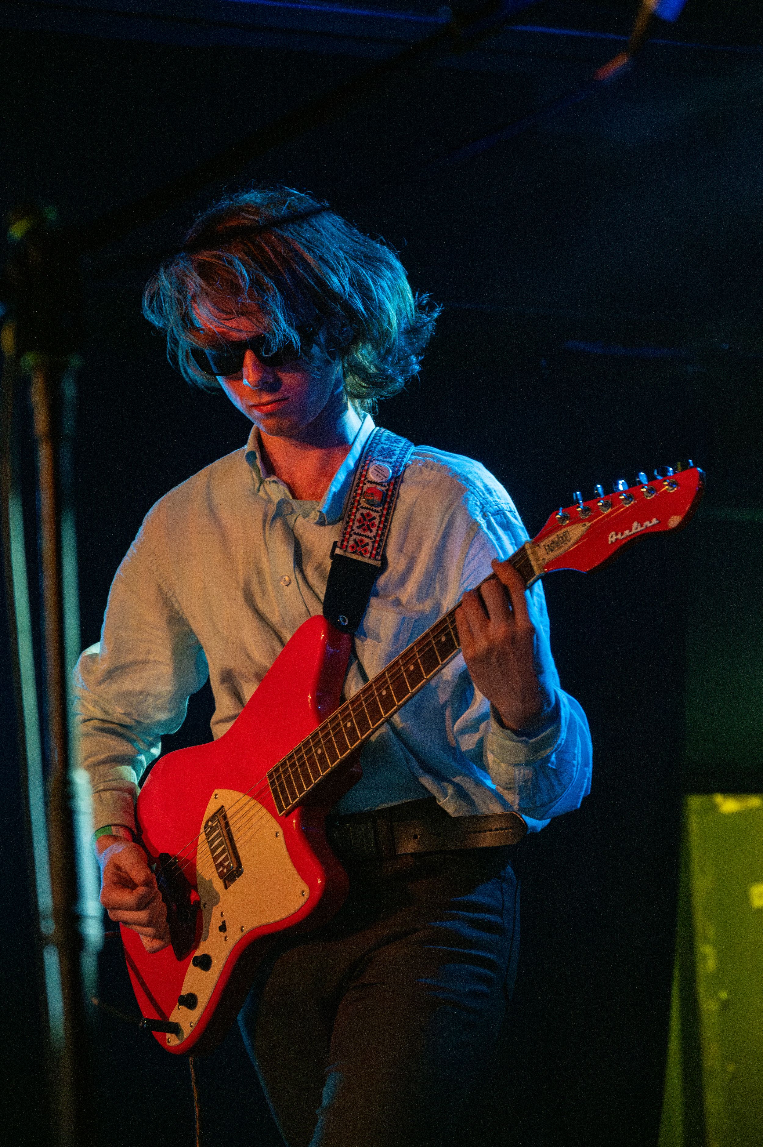 Musician plays red electric guitar under blue stage lighting while wearing white button-up shirt.