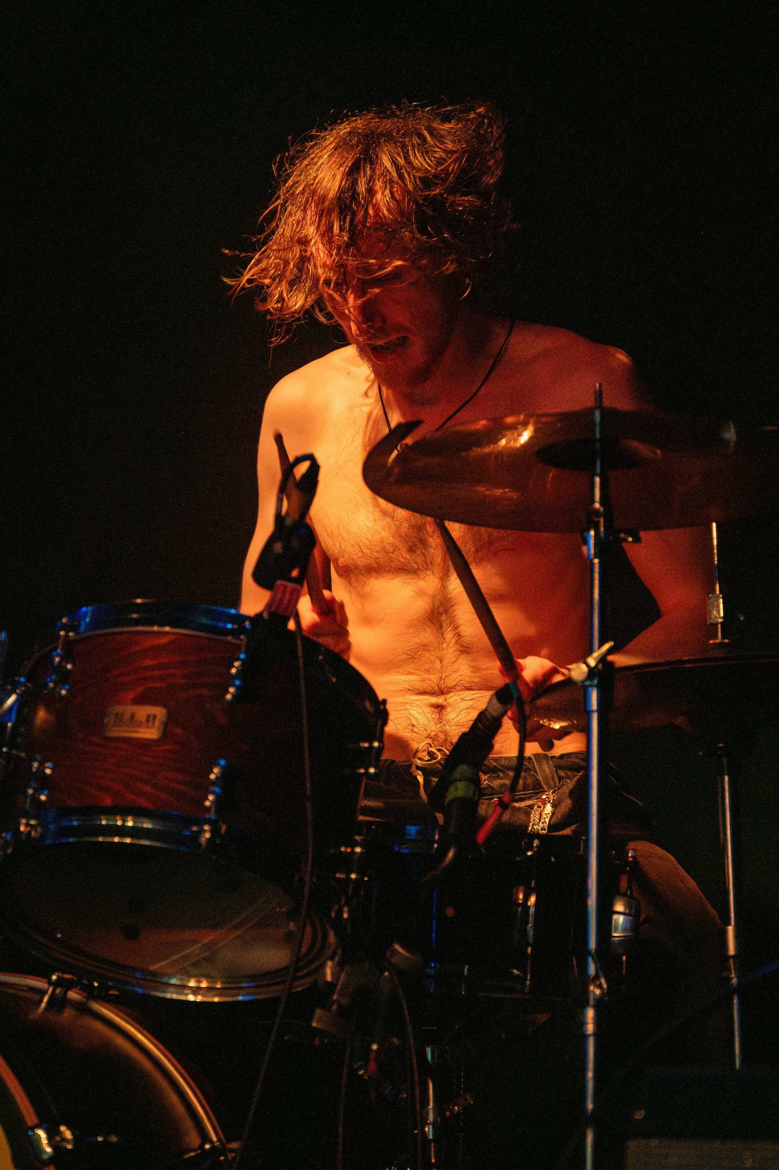 Shirtless drummer performs with intensity under warm orange stage lighting.