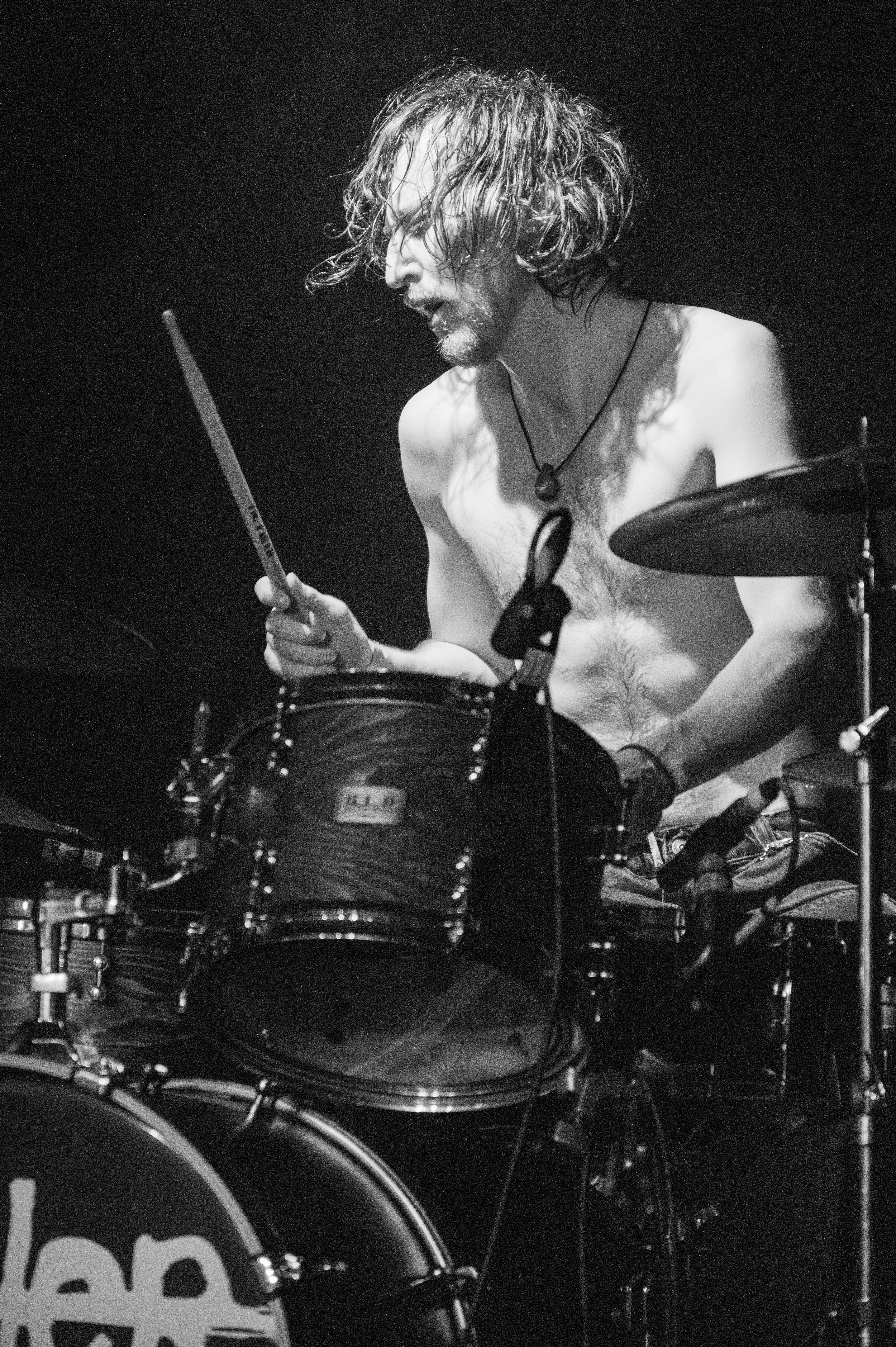 Black and white photo of shirtless drummer performing intensely behind drum kit on stage.