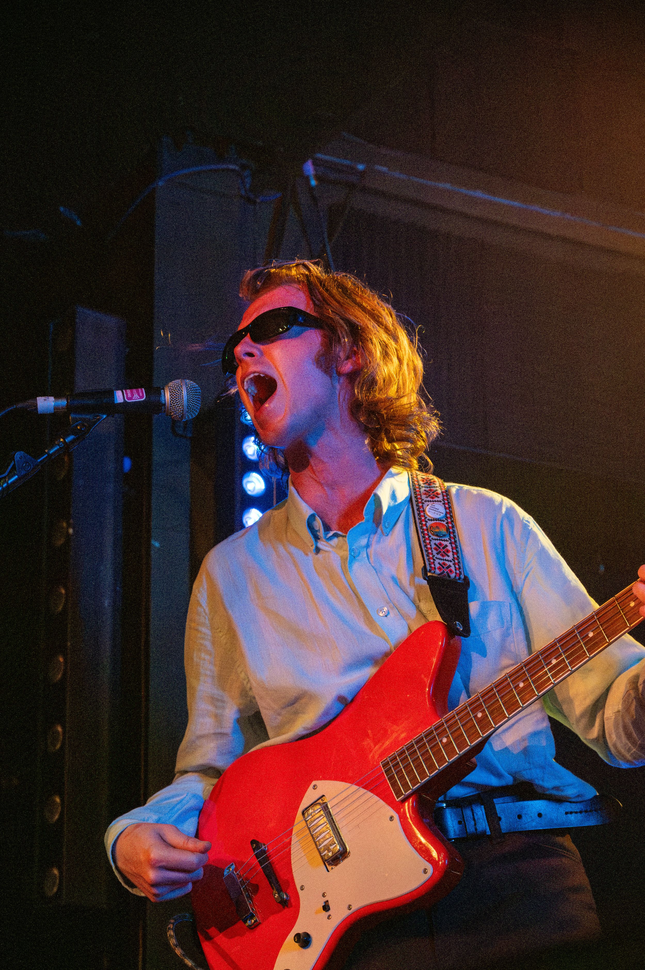Musician singing and playing red electric guitar while wearing sunglasses under blue stage lights.