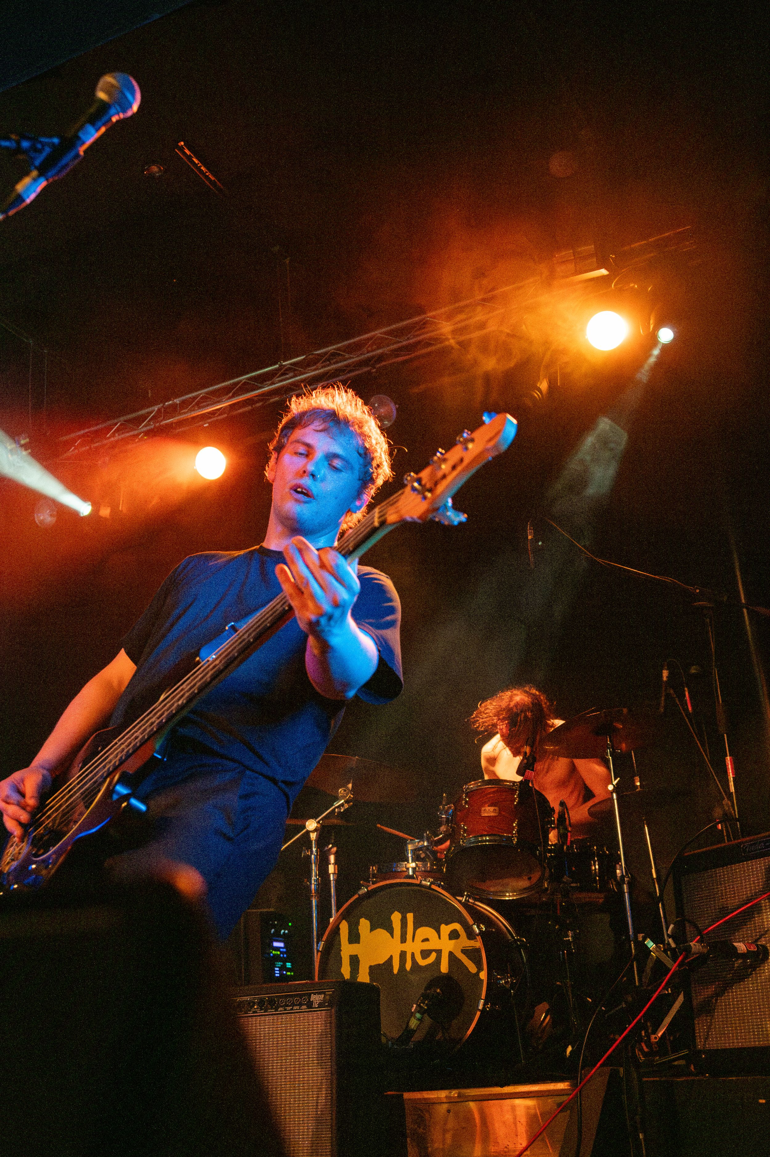 Musician plays bass guitar on stage with orange lighting and drum kit visible in the background.
