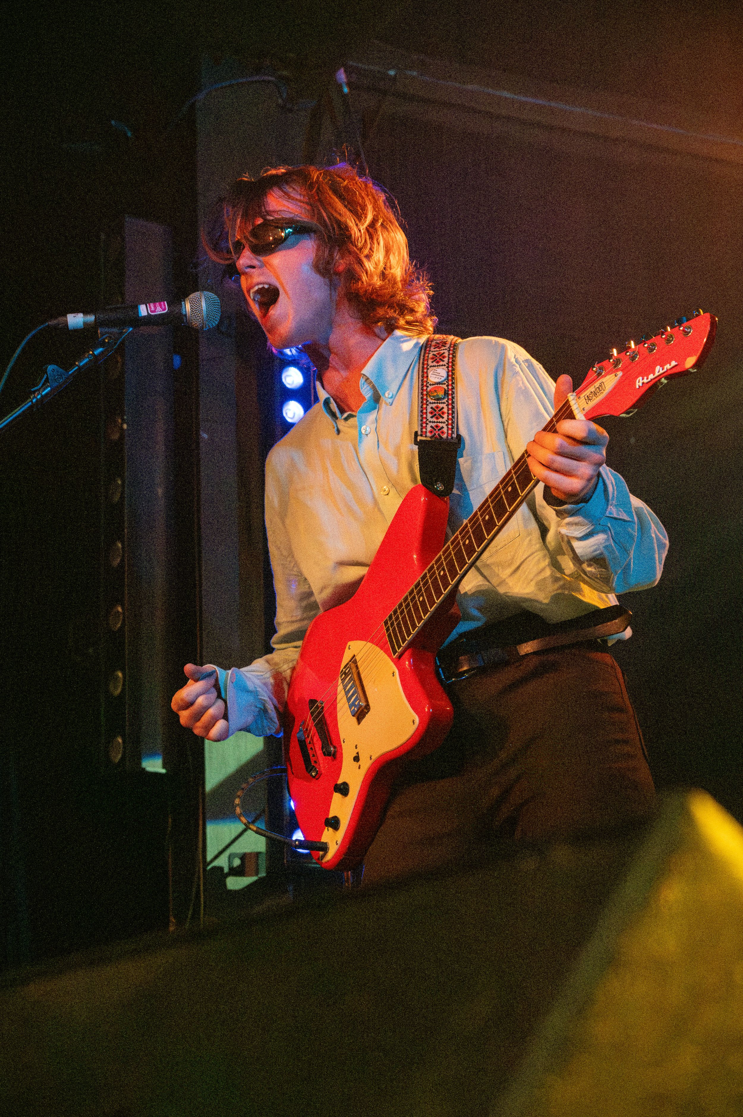 Musician in glasses plays red guitar while singing passionately on stage with bright lighting effects.