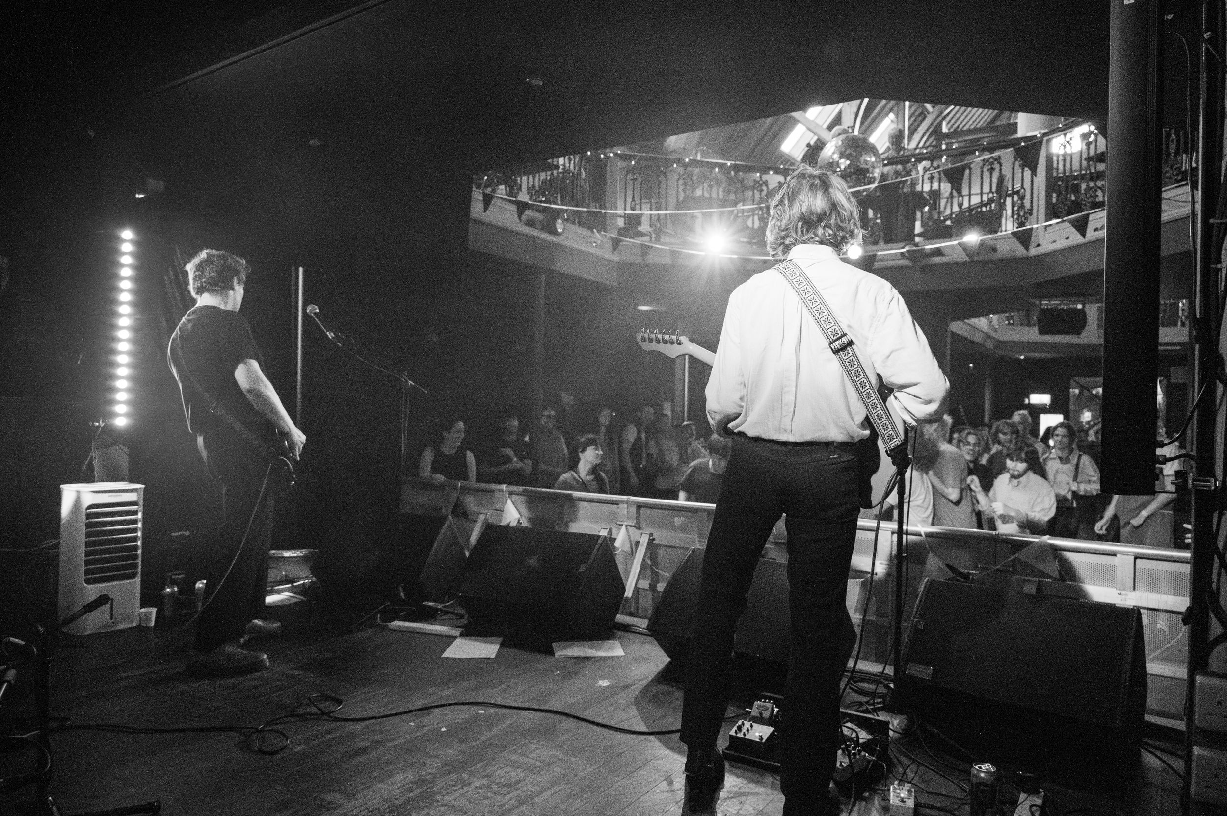 Black and white concert photo showing performers on stage with audience visible in the background.