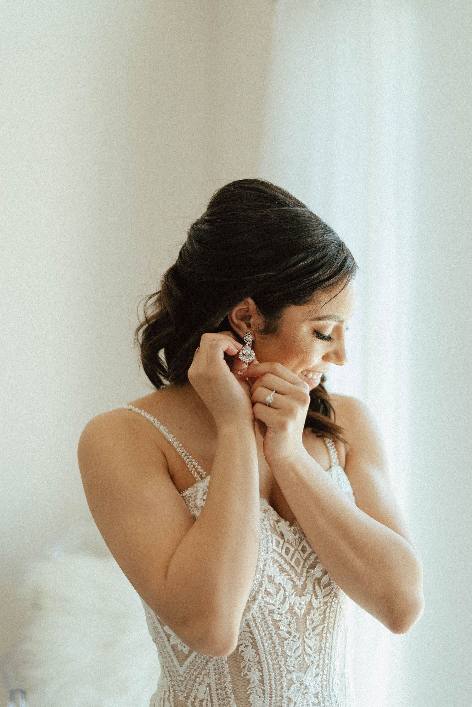 A bride in an intricately beaded wedding dress puts on earrings while getting ready.