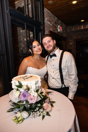 Newlyweds pose together by their wedding cake decorated with pink and purple flowers.