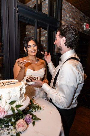 Bride and groom sharing cake together during reception with floral decorations on table. NJ wedding photographer
