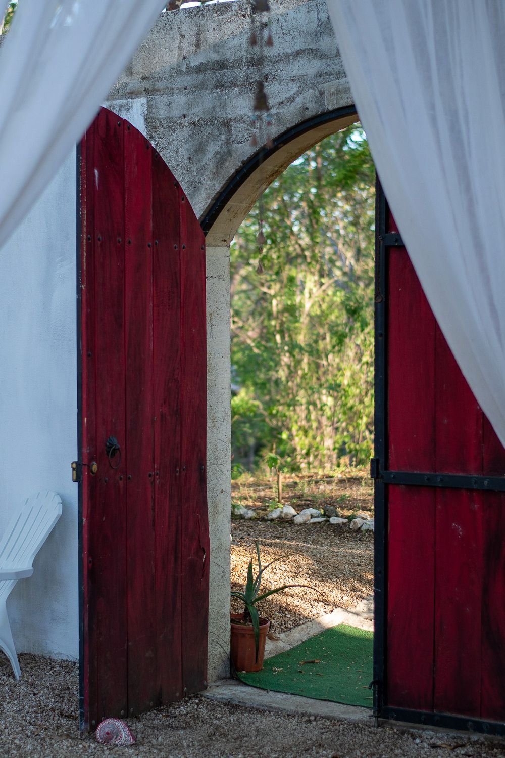 Red wooden doors with white curtains frame an archway leading to a sunlit garden.